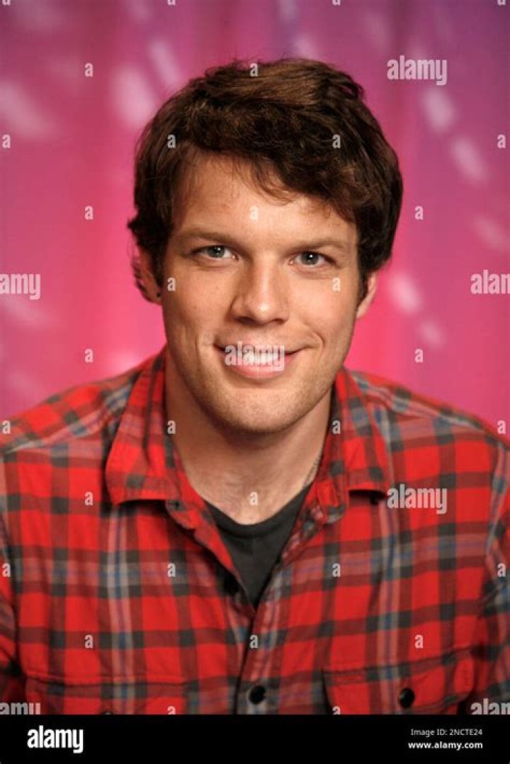 Jake Lacy posing in a white shirt and black suit.